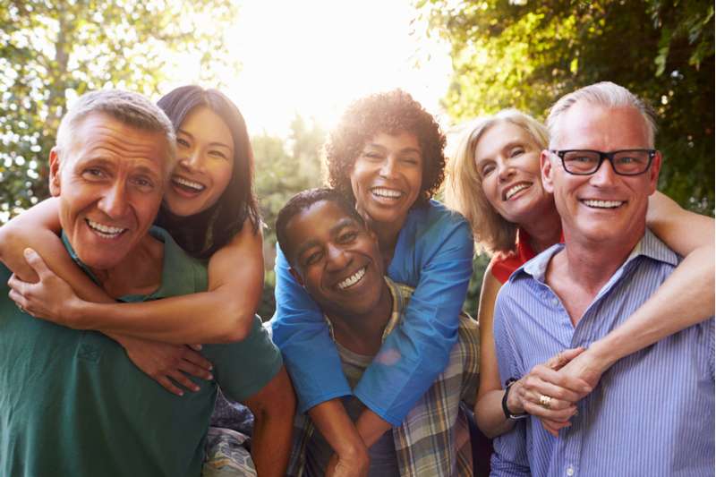 A group of six adults smiling and posing together outdoors in a sunny park, embodying the joy and connection often nurtured through marriage or couples counseling. Two individuals are giving piggyback rides, with everyone appearing happy and relaxed, surrounded by greenery. Rosen Couples Counselling