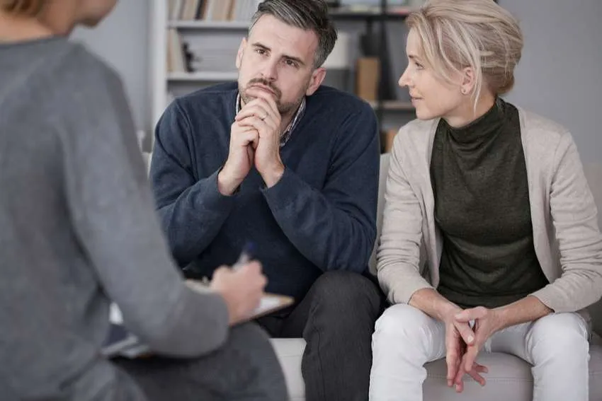 A man and woman sit on a couch, listening intently to a third person whose back is to the camera. The setting suggests marriage counselling or couples therapy, with bookshelves visible in the background containing various resources for relationship guidance and conflict resolution. Rosen Couples Counselling