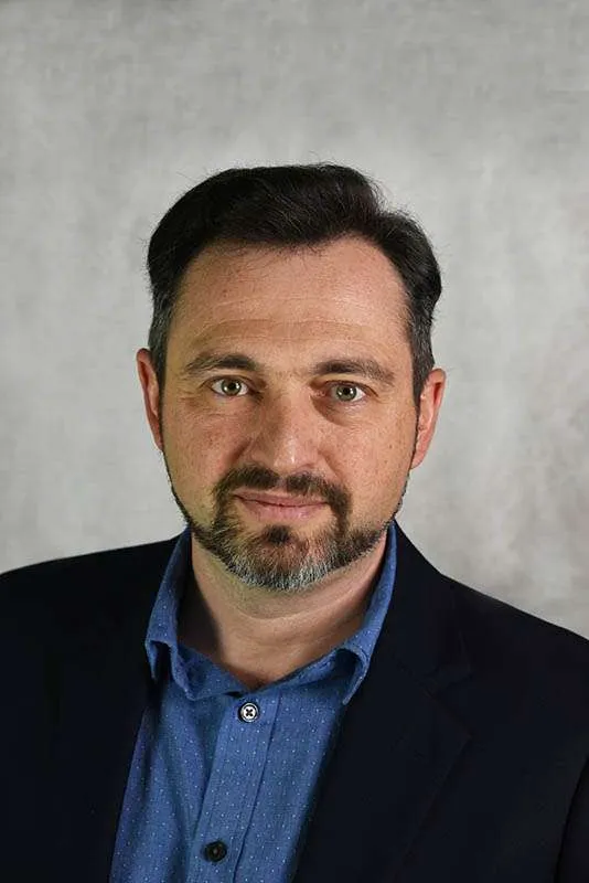 A man with short dark hair and a beard is wearing a blue shirt and black blazer, symbolizing the importance of seeking support in marriage or couples counselling. He looks directly at the camera against a neutral gray background, reflecting on the potential for growth and understanding within relationships when guided by professional help. Rosen Couples Counselling