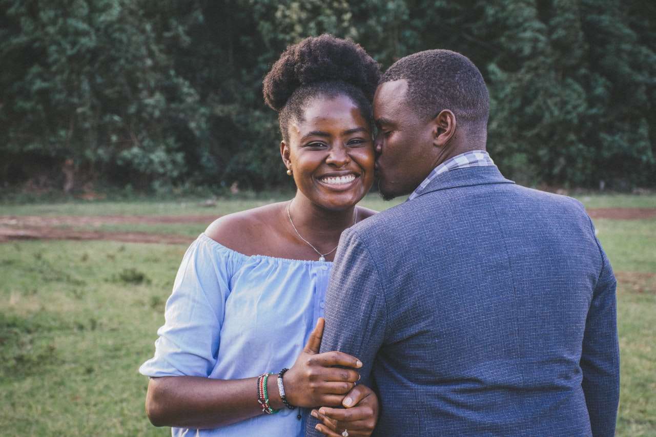 A couple stands outdoors; the woman smiles at the camera, wearing a light blue off-shoulder top, while the man in a gray jacket kisses her cheek. They are surrounded by greenery and appear happy and relaxed, embodying the tranquil joy often found through marriage and couples counselling. Rosen Couples Counselling