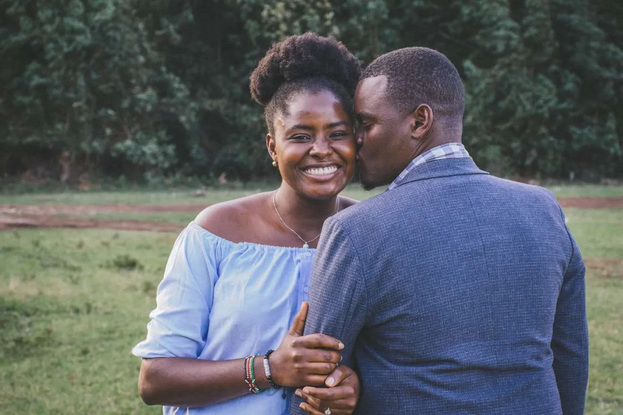 A couple stands outdoors; the woman smiles at the camera, wearing a light blue off-shoulder top, while the man in a gray jacket kisses her cheek. They are surrounded by greenery and appear happy and relaxed, embodying the tranquil joy often found through marriage and couples counselling. Rosen Couples Counselling