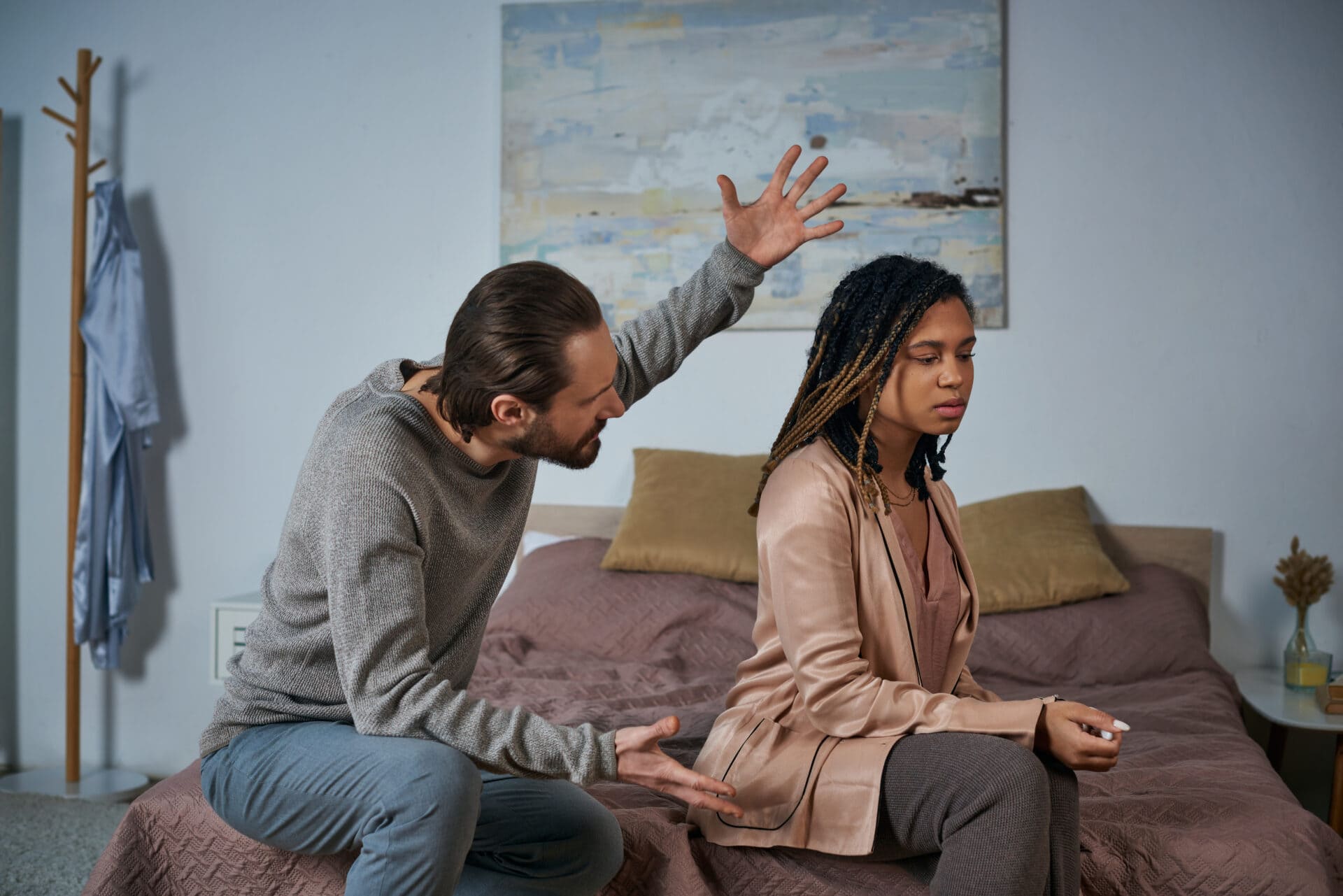 A man and woman sit on a bed in a tense situation, illustrating the challenges that lead to marriage counselling. The man gestures with his arms raised, embodying the chaos of anger management gone awry—a frequent issue addressed in couples counselling. The woman sits facing away, eyes closed, exuding calm detachment amidst the softly lit room, symbolizing her need for emotional distance and reflection often explored during relationship therapy sessions. Rosen Couples Counselling