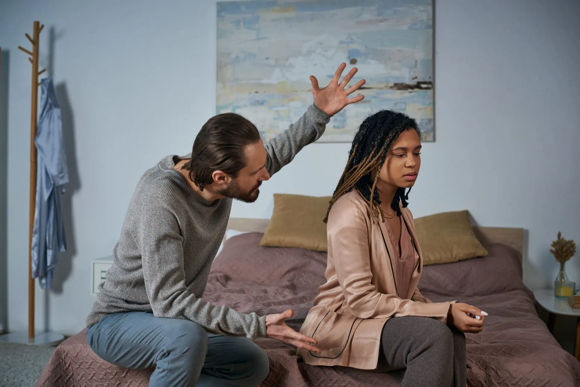 A man and woman sit on a bed in a tense situation, illustrating the challenges that lead to marriage counselling. The man gestures with his arms raised, embodying the chaos of anger management gone awry—a frequent issue addressed in couples counselling. The woman sits facing away, eyes closed, exuding calm detachment amidst the softly lit room, symbolizing her need for emotional distance and reflection often explored during relationship therapy sessions. Rosen Couples Counselling