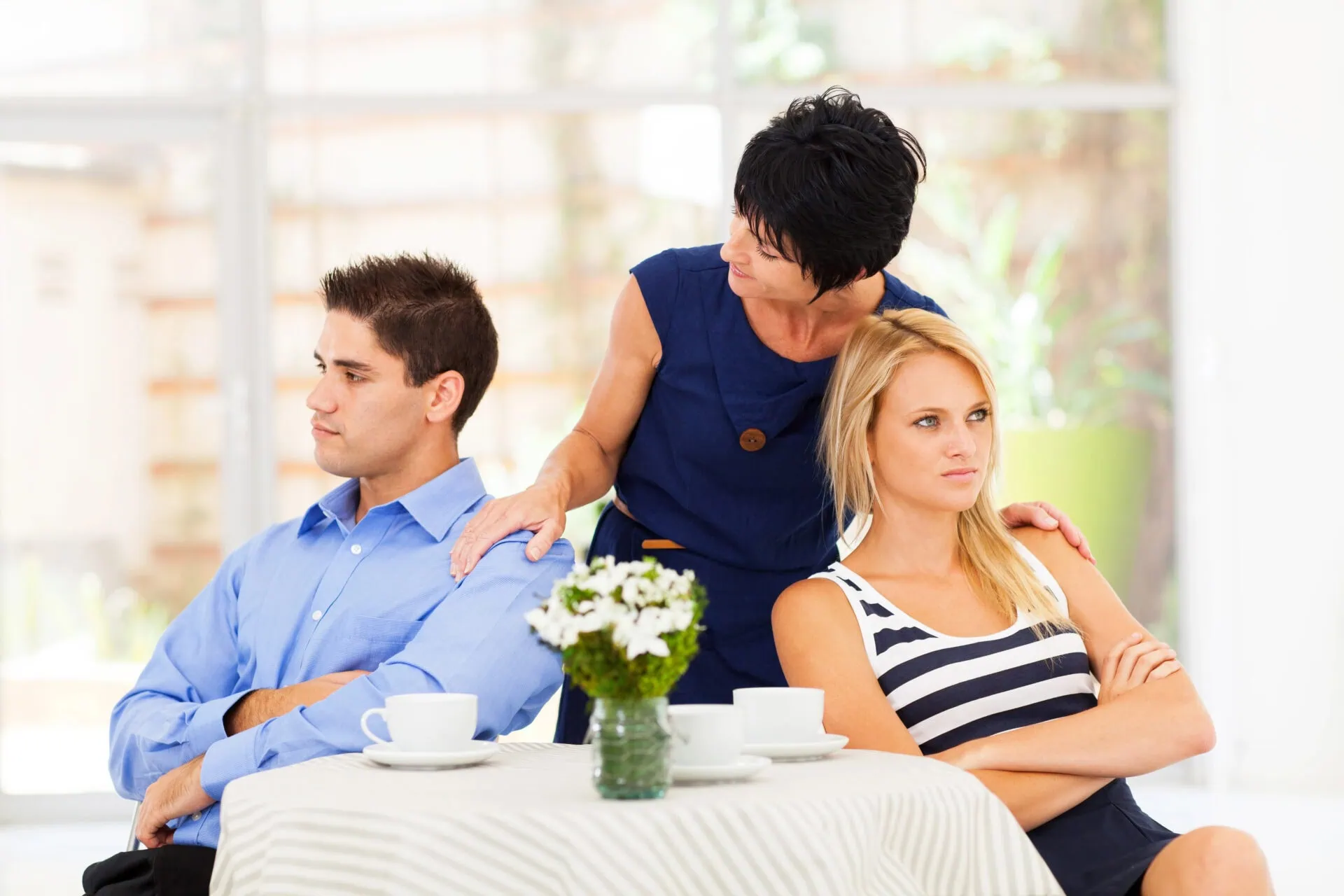 A woman stands between a man and a woman seated at a table, arms crossed in frustration. The couple avoids eye contact, perhaps reflecting on the challenges of marriage counselling and the potential impact on their relationship. A small vase with white flowers and white cups decorate the bright and airy setting, symbolizing hope amidst tension. Rosen Couples Counselling