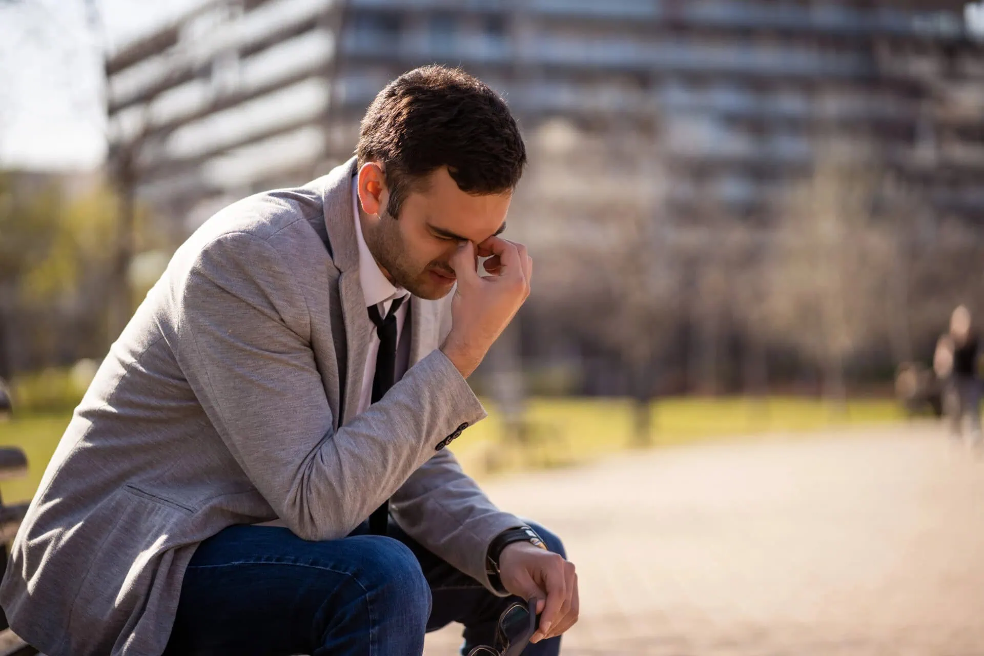 A man in a gray blazer and jeans sits outdoors, looking stressed as he struggles to express his feelings, pinching the bridge of his nose. This moment reflects the challenges often addressed in marriage counseling or couples counseling. In the background, a modern building and trees are blurred under the sunlight, casting calming shadows on the pavement, symbolizing hope for resolution and understanding through therapy. Rosen Couples Counselling