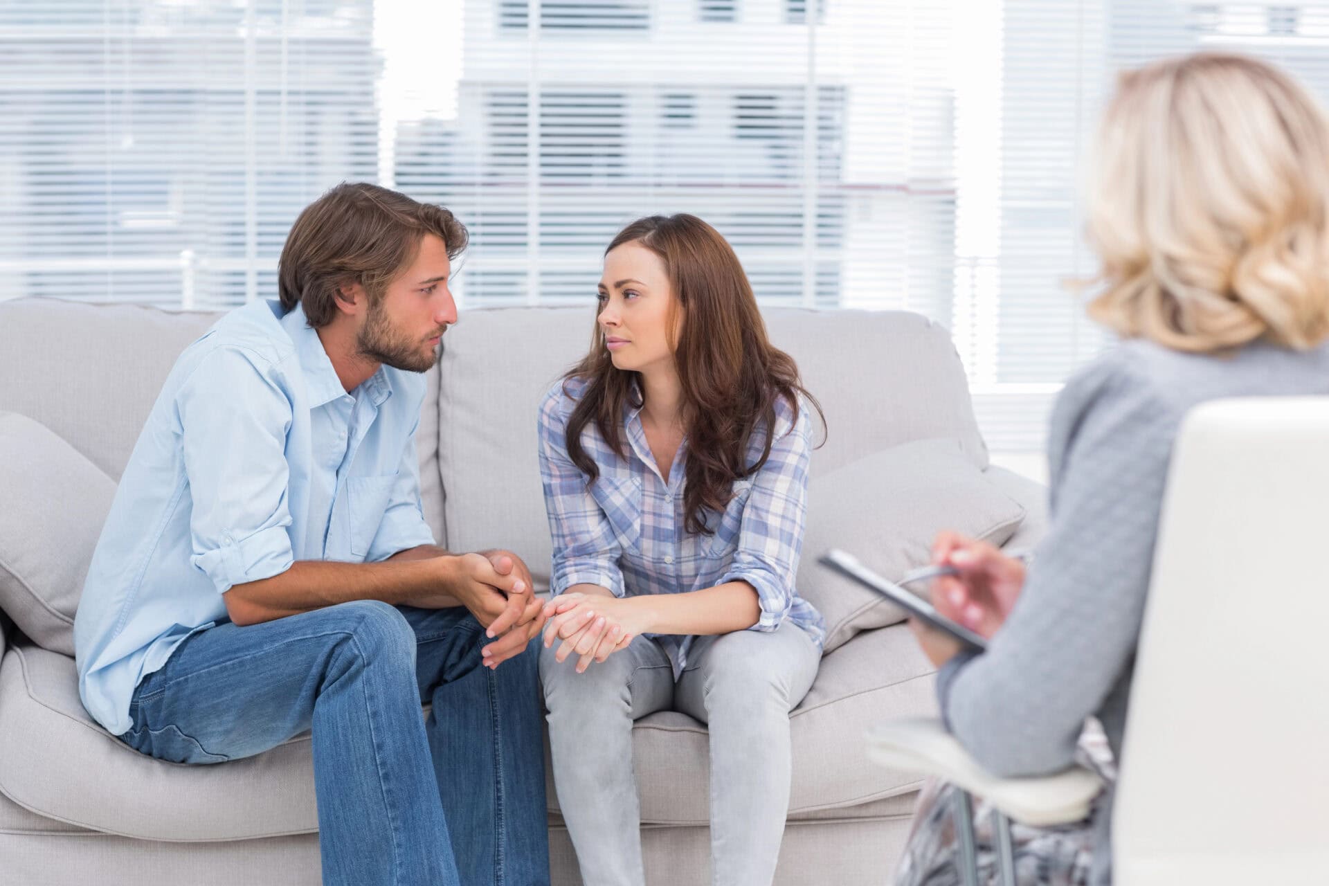 A couple sits on a sofa, facing each other, holding hands. A therapist with a notepad is seated opposite them, guiding the session as part of couples therapy or marriage counselling. The room’s large windows with blinds create a bright and open atmosphere. Rosen Couples Counselling
