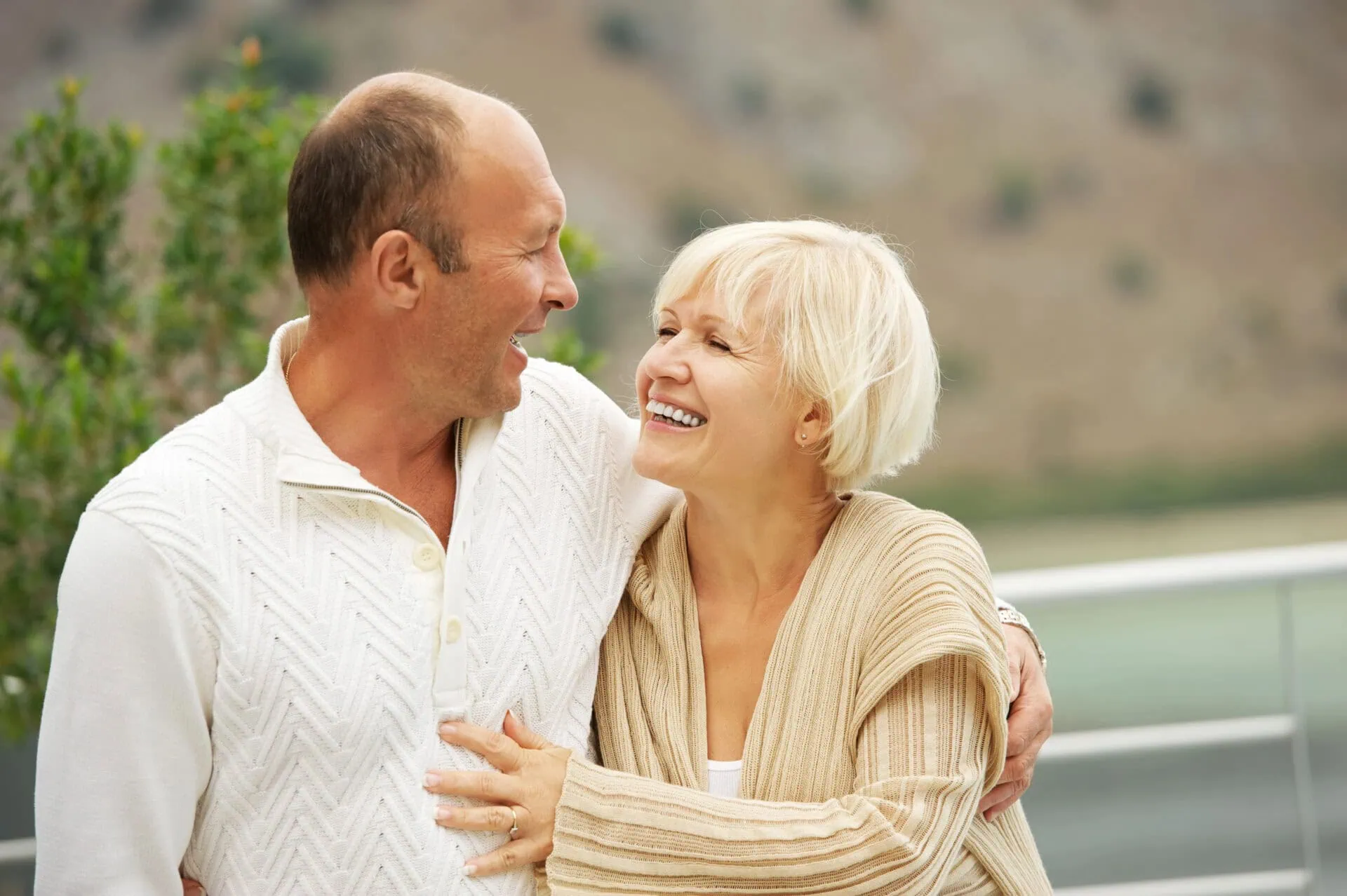An older couple is outdoors, smiling and embracing each other joyfully. They are standing near a railing with a blurred natural landscape in the background, illustrating how effective marriage counseling can help overcome common relationship challenges for lasting happiness. Both are wearing light-colored sweaters. Rosen Couples Counselling