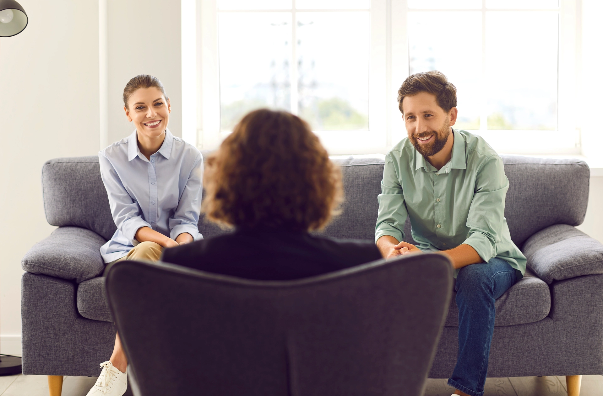 Young family couple visiting professional therapist or psychologist. Smiling, cheerful husband and wife sitting on sofa and having therapy session together. Back view of psychotherapist in armchair
